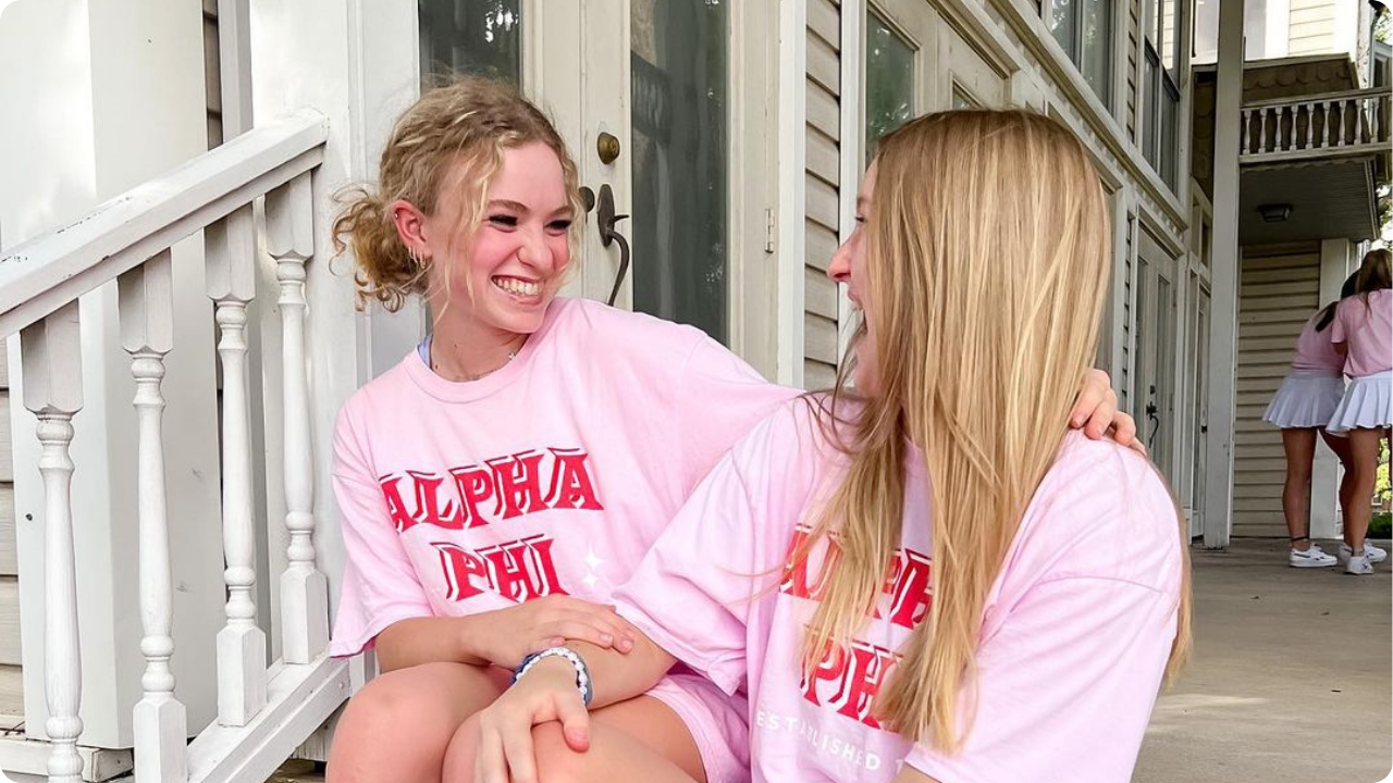 Two girls wearing pink Alpha Phi shirts while sitting on porch stairs
